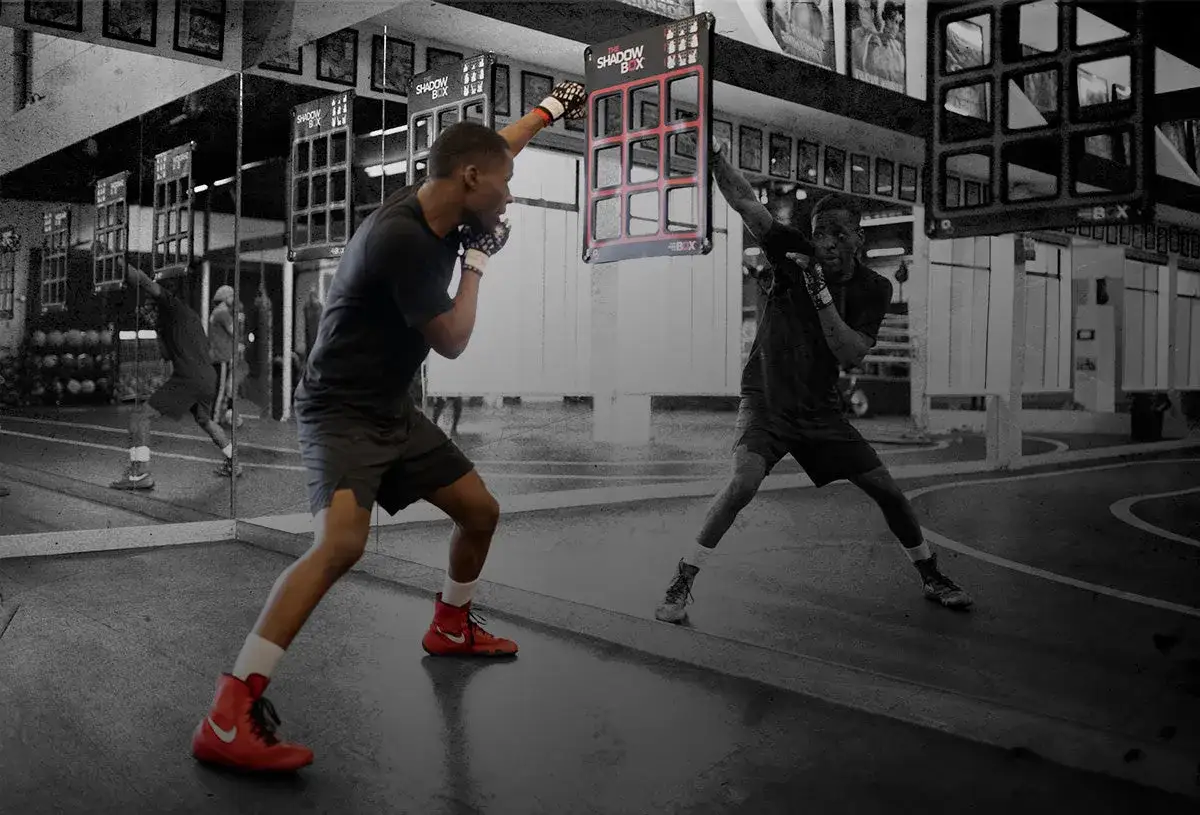 Two men practice shadow boxing in a gym, one striking a target board. This shows the shadow boxing trend in fitness.