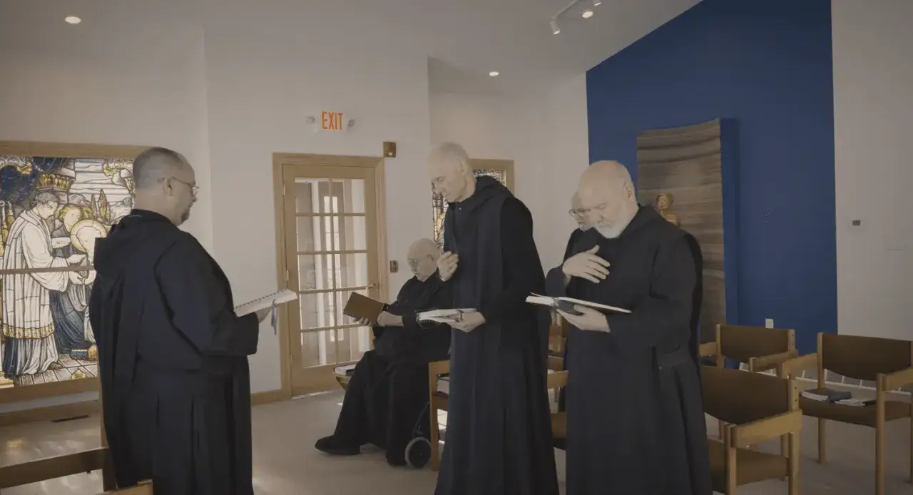 Four monk priests in dark robes stand and sit, reading from books in a bright room with stained glass.