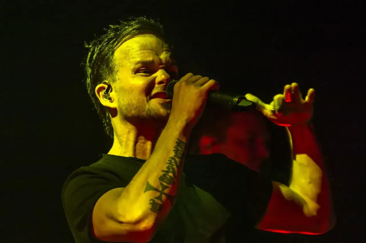 The Rasmus wokalista, z tatuażem na ramieniu, śpiewa do mikrofonu, oświetlony żółtym i czerwonym światłem.