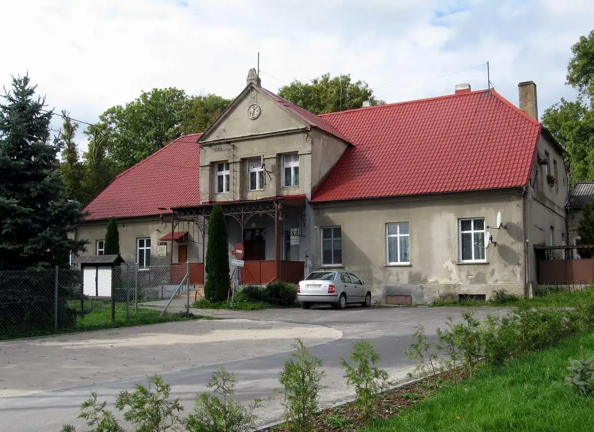 Budynek z czerwonym dachem, przypominający trochę dom z bajki o robakach, stoi pośród zieleni. Obok zaparkowany srebrny samochód.