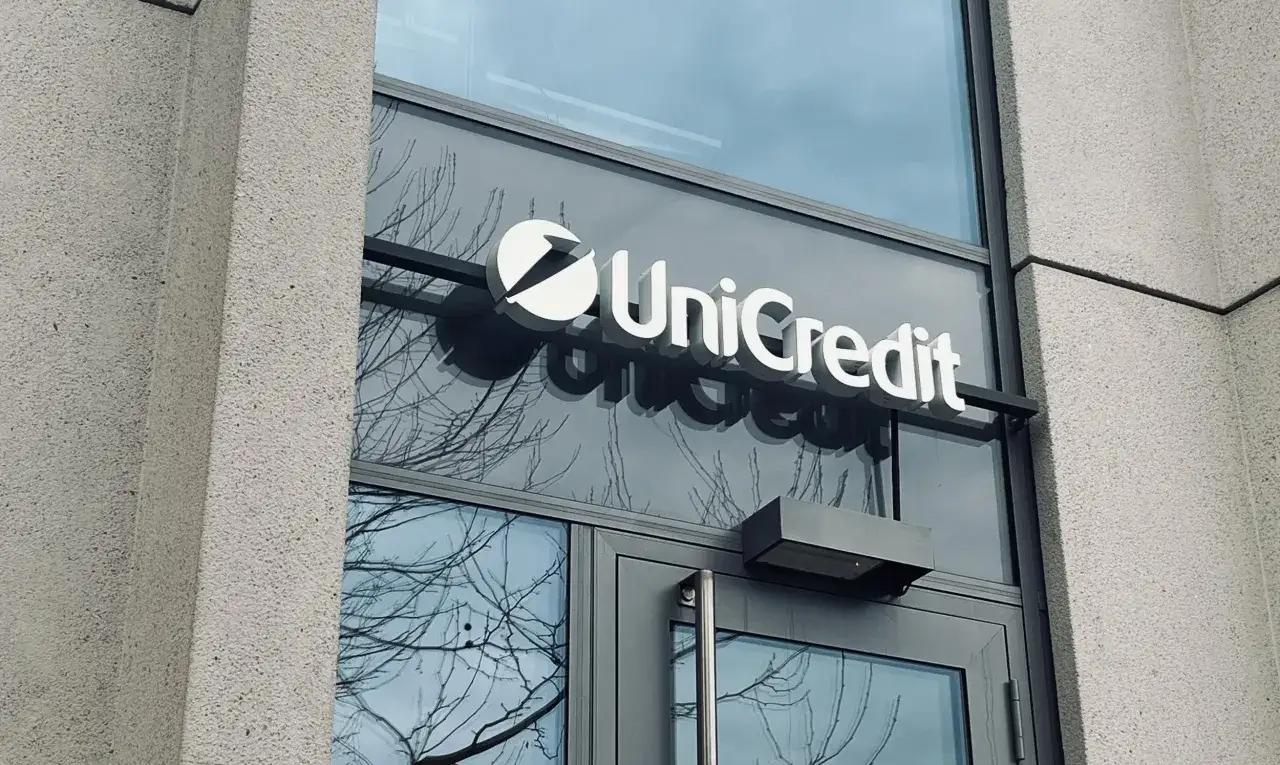 Logo banku UniCredit na szklanych drzwiach. W tle odbijają się nagie gałęzie drzew.