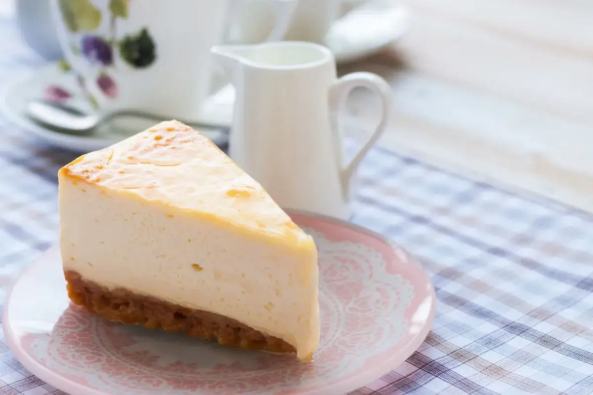 Una fetta di cheesecake con mascarpone, su un piattino rosa, con una tazza da tè e una lattiera sullo sfondo.