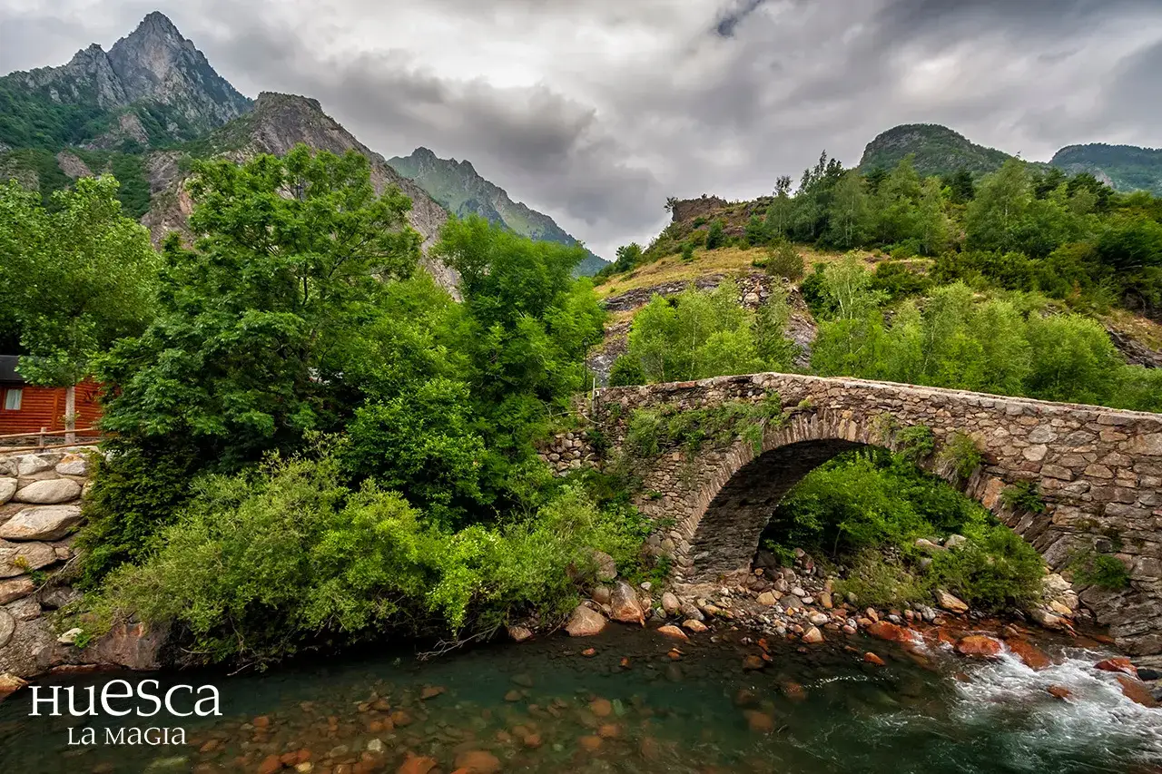 Puente de piedra sobre un río en el **valle de Vallibierna**, rodeado de exuberante vegetación y montañas imponentes bajo un cielo nublado.