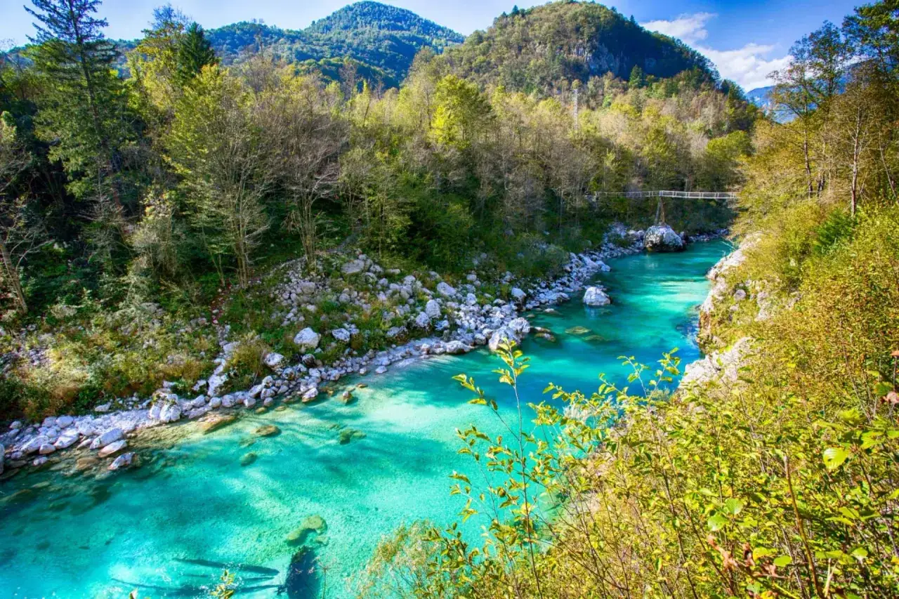 Vandra i Slovenien l&auml;ngs en turkos flod kantad av stenar och gr&ouml;nskande berg. En bro syns i fj&auml;rran.
