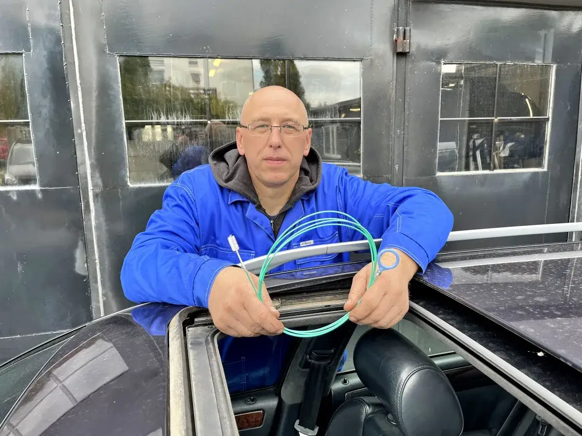 Mechanik w niebieskim kombinezonie czyści otwory odpływowe w samochodzie za pomocą zielonego wężyka.
