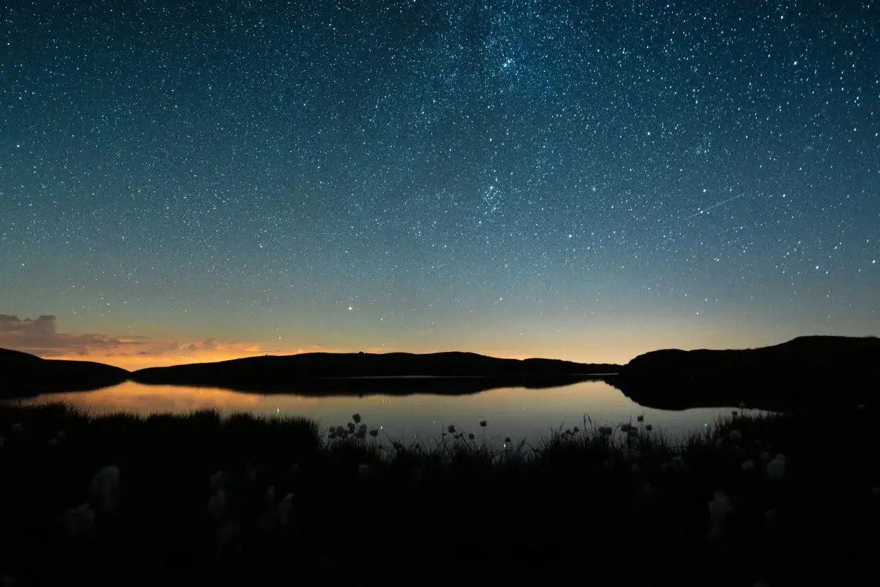 Nocne niebo pełne gwiazd nad jeziorem. W oddali widać pomarańczowy blask zachodzącego słońca. Gwiazda polarna inaczej, jako część tego kosmicznego spektaklu.