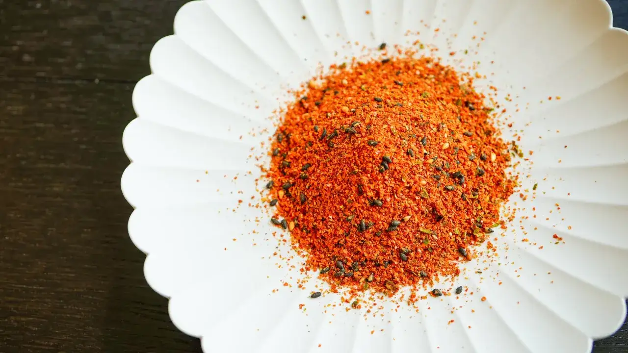 A pile of vibrant Japanese 7 spice blend, featuring chili flakes and black sesame seeds, rests on a white, fluted dish.