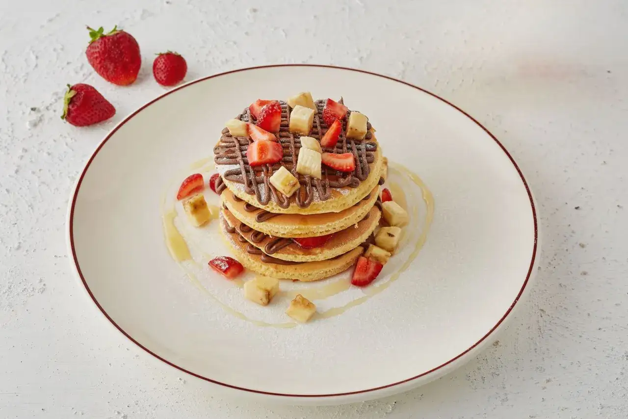 Pancake senza latte con crema di nocciole, fragole e banane a cubetti, guarniti con sciroppo d'acero e zucchero a velo.