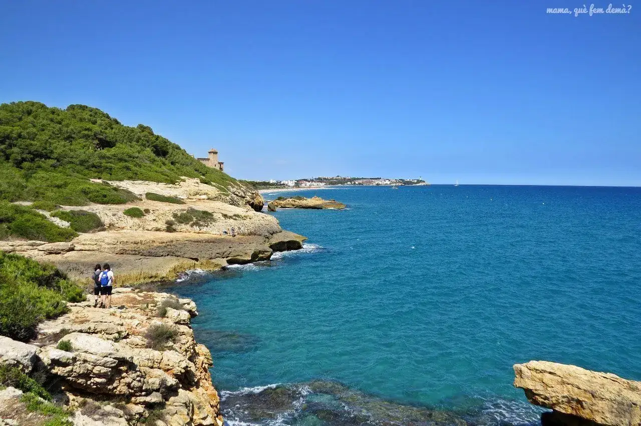 Dos personas caminan por el cam&iacute; de ronda de Tarragona, disfrutando de las vistas al mar azul y la costa rocosa.