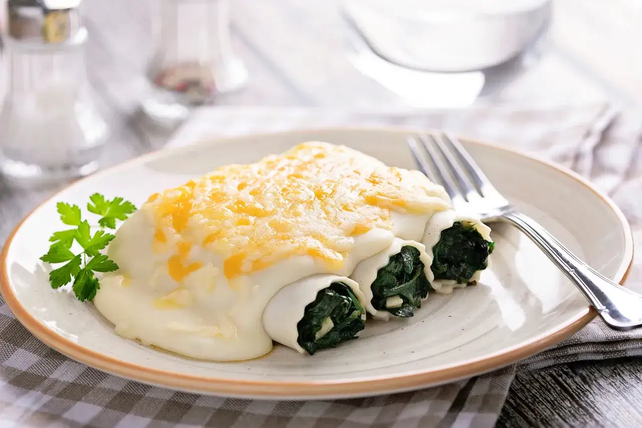 Canelones de espinacas cubiertos de bechamel y queso gratinado, servidos en un plato con perejil fresco.