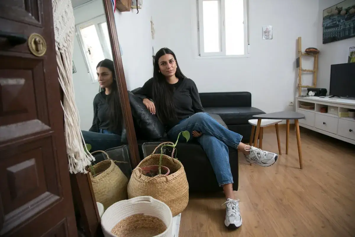 Joven sentada en sofá negro, reflexiona sobre el bono joven alquiler. Cestas de mimbre y planta en primer plano.