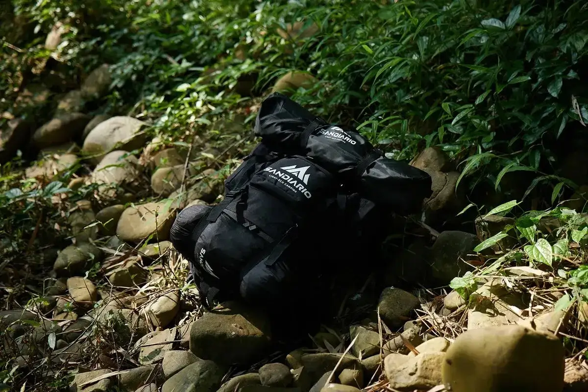Mochila negra lista para el bushcraft, con equipo de camping en un entorno natural rocoso y frondoso.