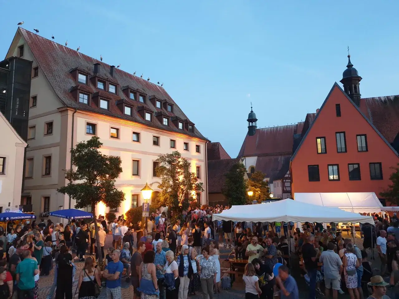 Menschen feiern bei Abendveranstaltungen in Herrieden. St&ouml;rche sitzen auf den D&auml;chern historischer Geb&auml;ude.