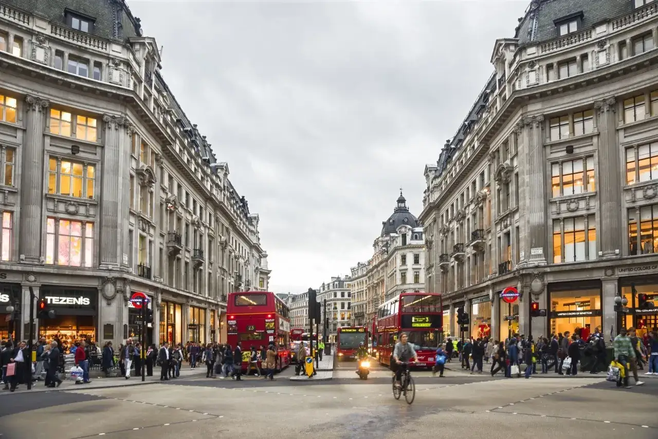 Trafik och folk på en livlig gata med klassiska byggnader, typiska london butiker och röda dubbeldäckare.