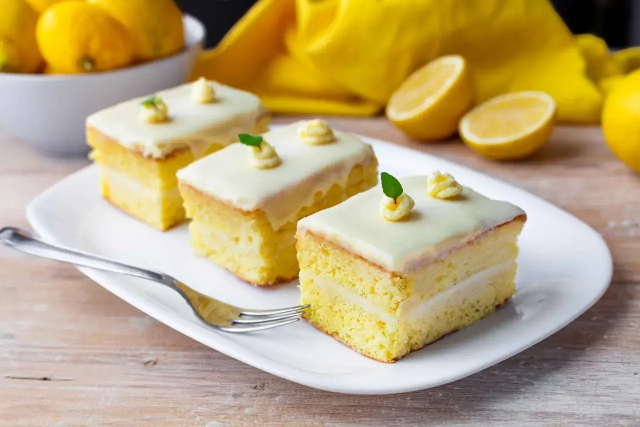 Tre fette di torta al cioccolato al limone, ricoperte di glassa bianca e guarnite con ciuffi di crema e foglioline verdi, su un piatto bianco con una forchetta.
