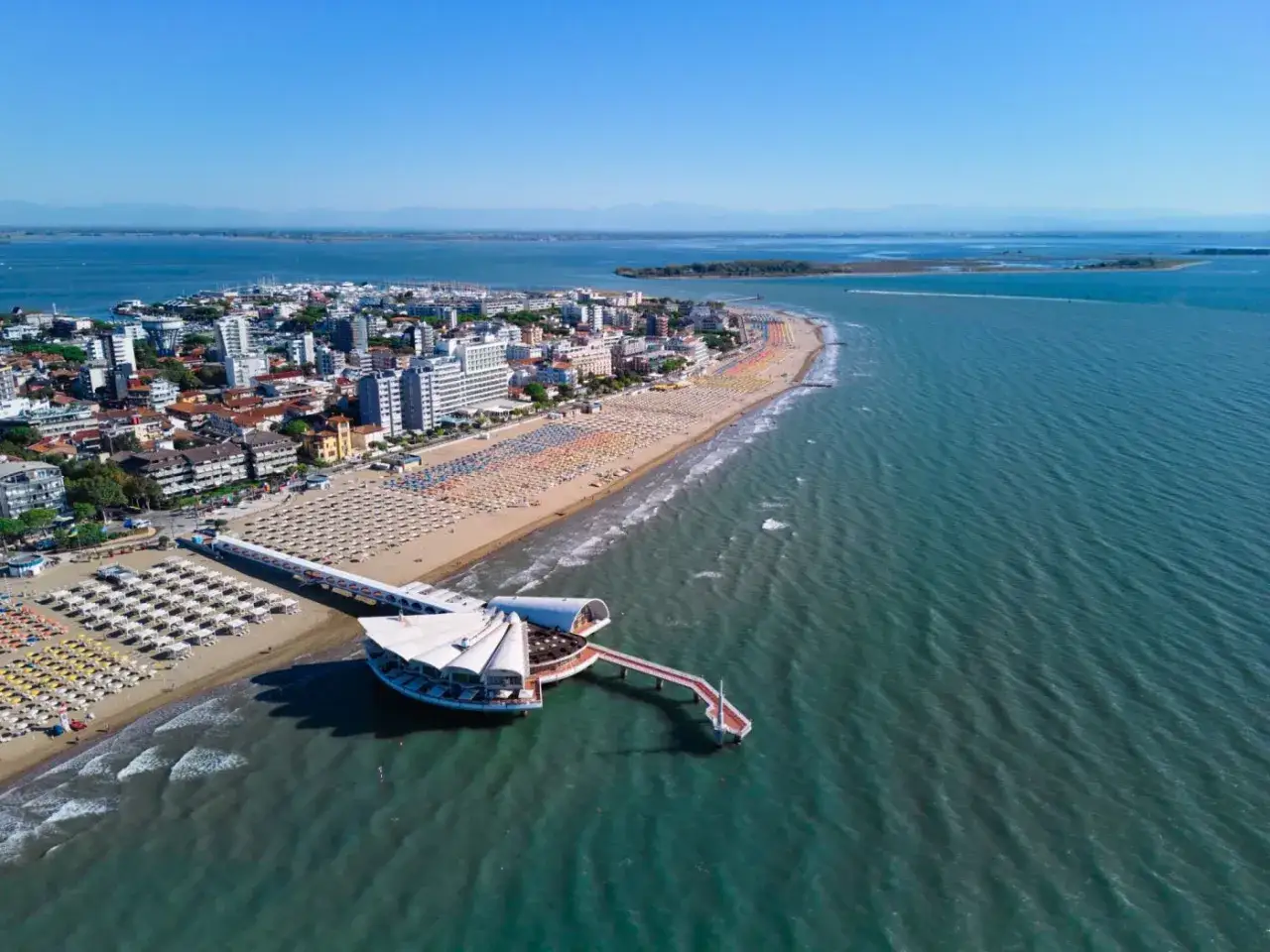 Vista aerea di una vivace località balneare con file di ombrelloni colorati sulle spiagge friulane. Un moderno pontile si estende sul mare azzurro.