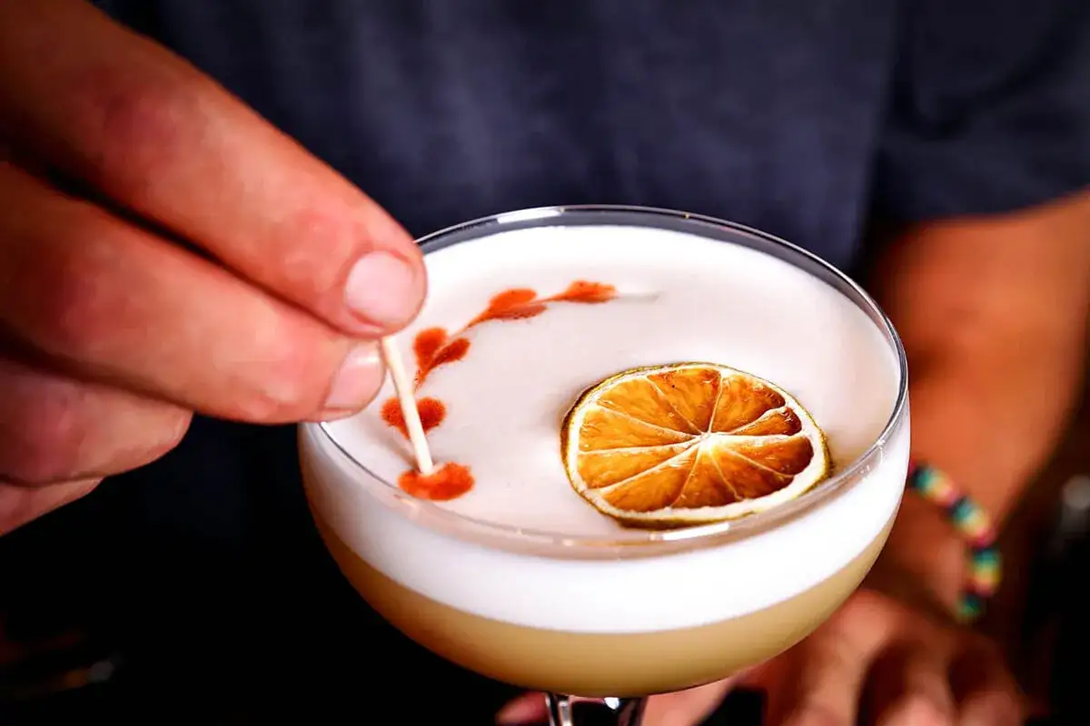En hand dekorerar en perfekt whisky sour med en tunn pinne, en torkad lime och ett mönster av bitters.