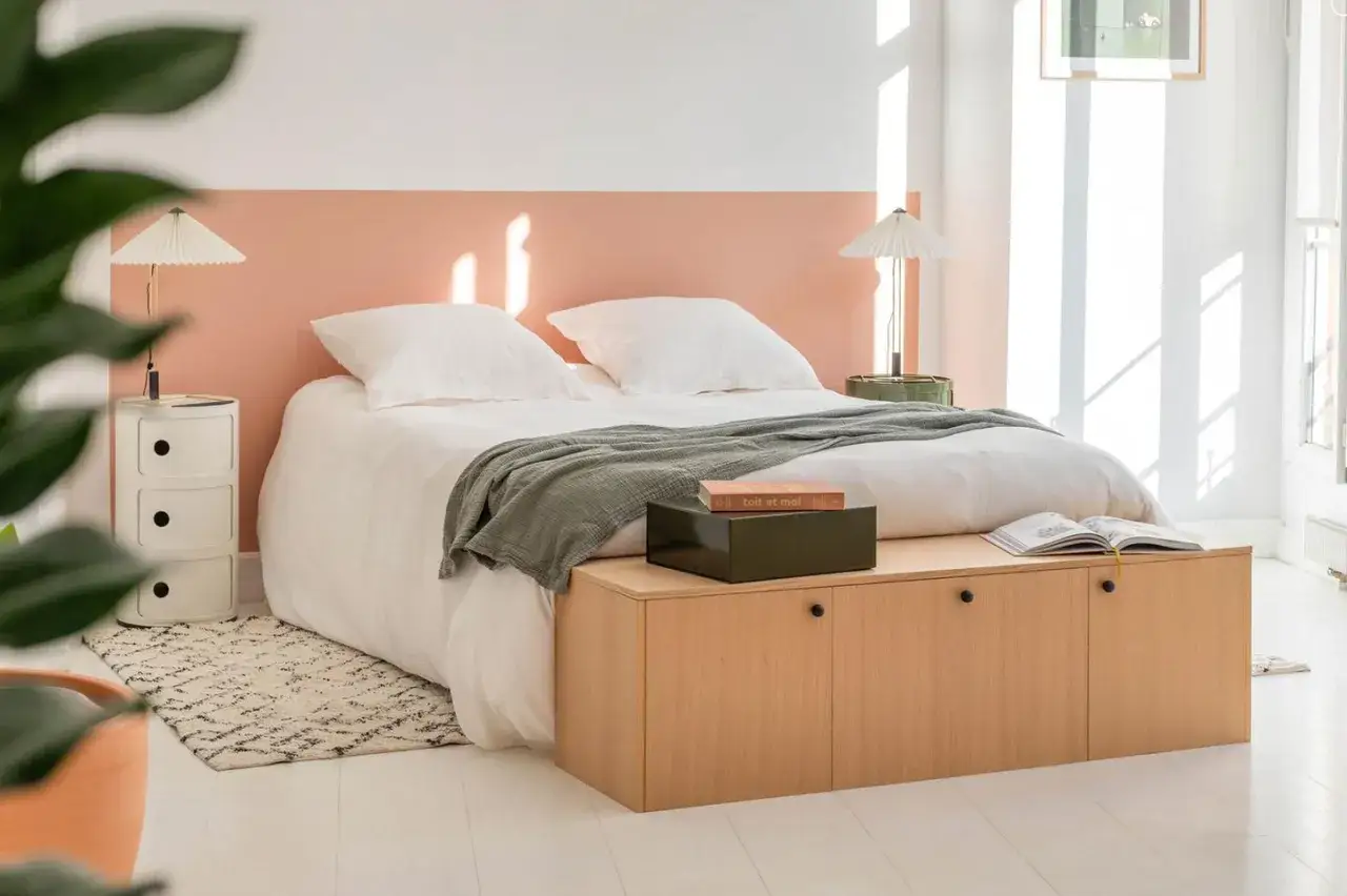 Schlafzimmer mit weißem Bett, Laken und grüner Decke. Die Wand nur teilweise streichen in zartem Rosa. Holzbank mit Büchern.