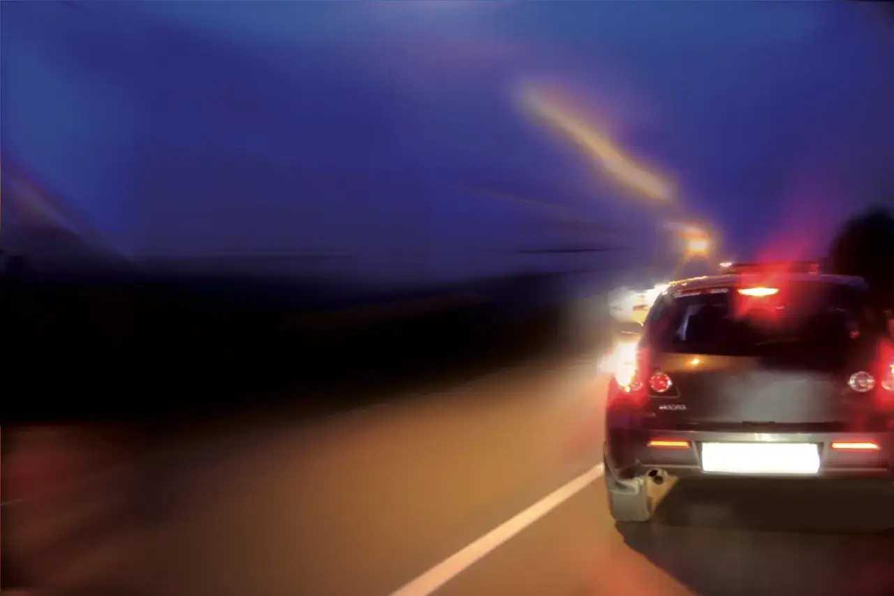 Un coche oscuro con luces traseras rojas y una luz de freno encendida, conduciendo de noche por una carretera vacía.