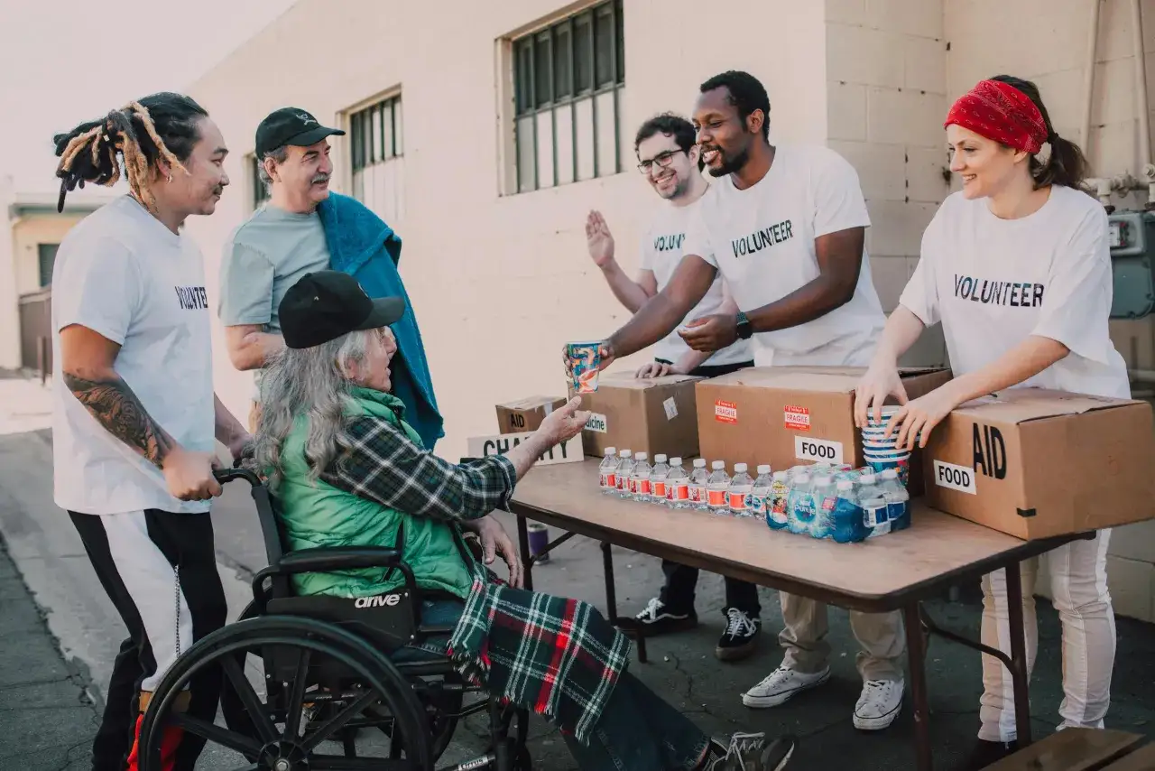 Volunteers in white t-shirts distribute food and aid. A person in a wheelchair receives a cup from a volunteer, showcasing community support.