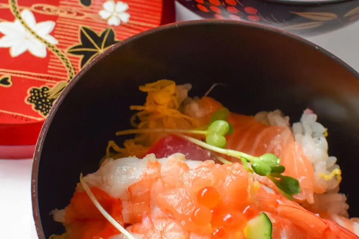 Chirashi zushi z krewetkami, łososiem i ikrą łososia, podane w czarnej misce, obok ozdobnego czerwonego pudełka.