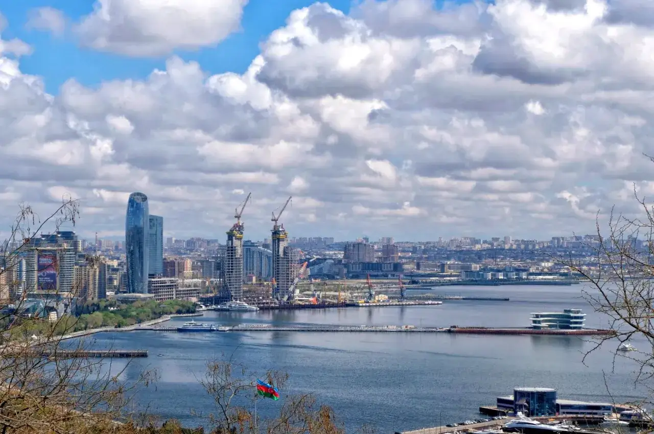Widok na Baku z morzem i chmurami. Widać nowoczesne budynki, dźwigi i flagę Azerbejdżanu. Baku pogoda zapowiada się słonecznie.