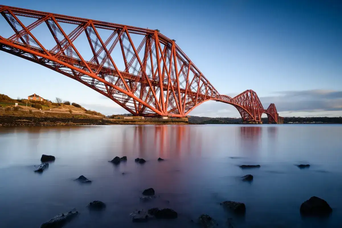 El icónico puente de Forth se alza majestuoso sobre aguas tranquilas, reflejando su estructura de hierro rojo contra un cielo azul claro.