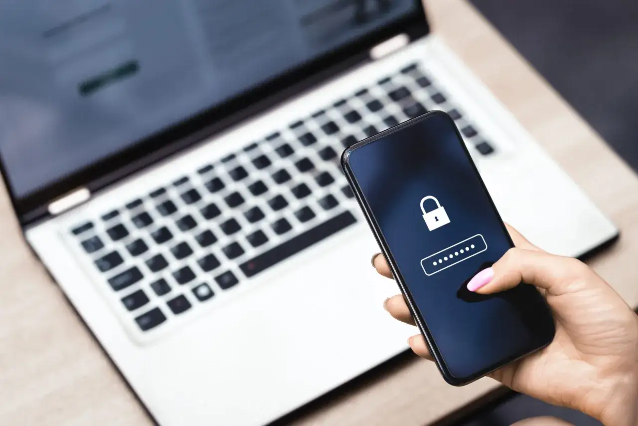 Dłoń z różowym lakierem na paznokciach trzyma smartfon z ikoną kłódki i polami do wpisania hasła. Obok leży laptop. To dziwne telefony, które wymagają uwierzytelnienia.