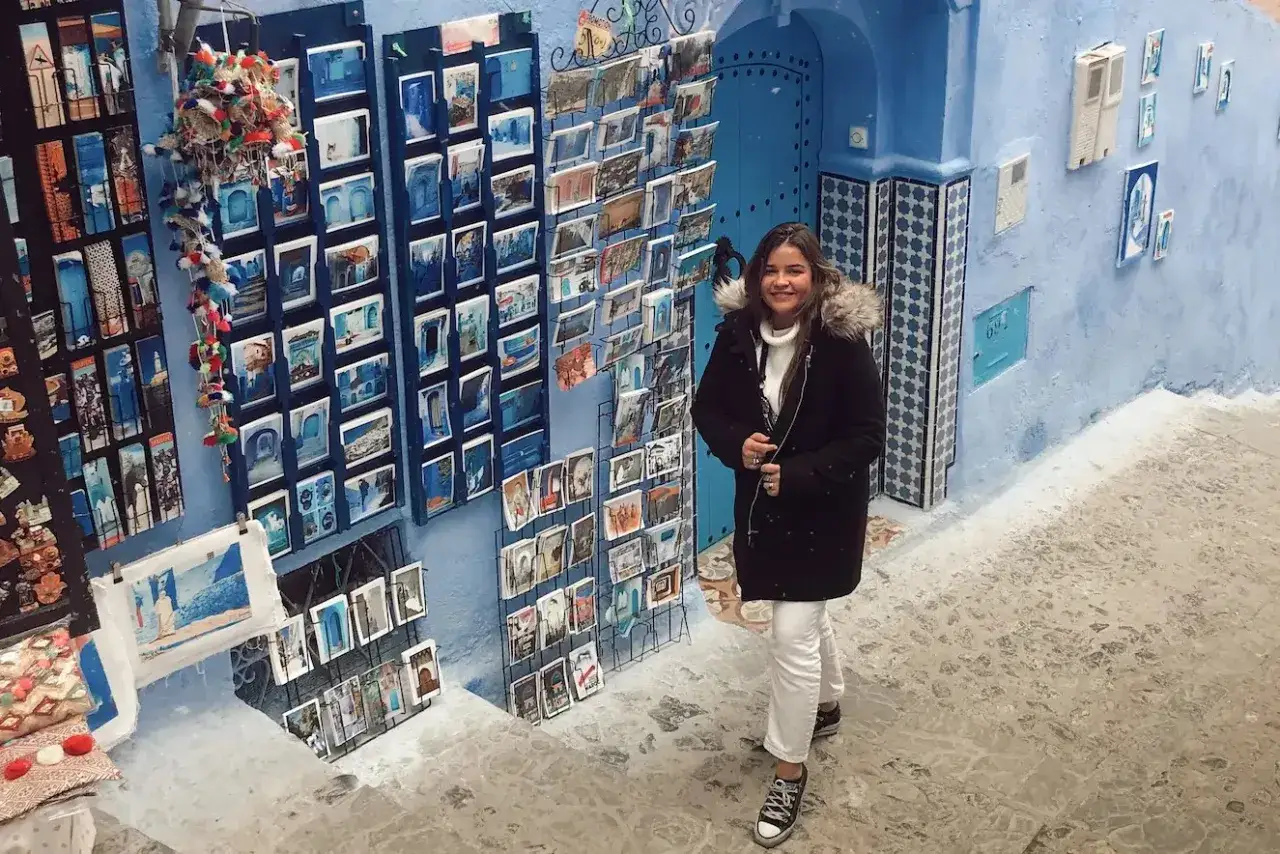 Una mujer sonríe frente a una pared azul llena de postales en Marruecos. Viajar a Marruecos para mujeres es una experiencia segura y enriquecedora.