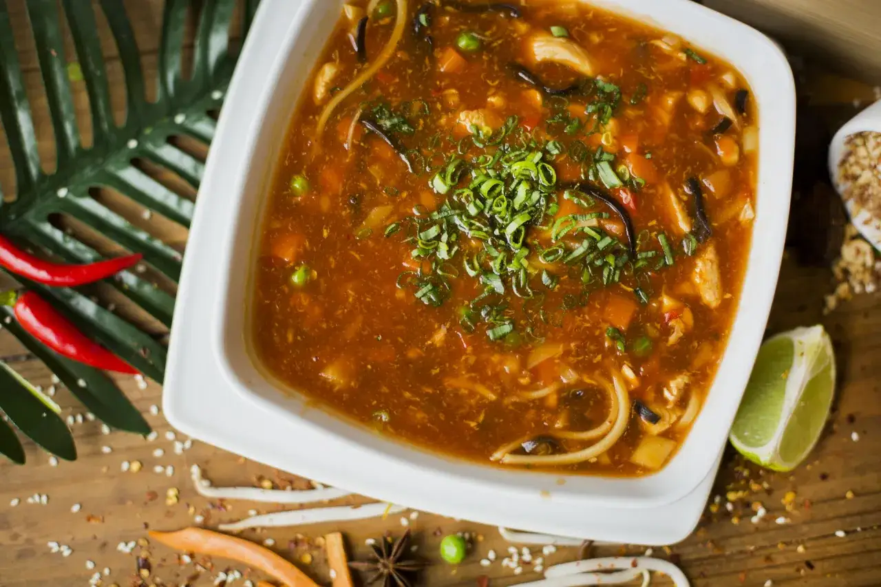 Aromaticzna zupa po pekińsku z kawałkami kurczaka, warzywami i makaronem, posypana szczypiorkiem. Obok chili i limonka.