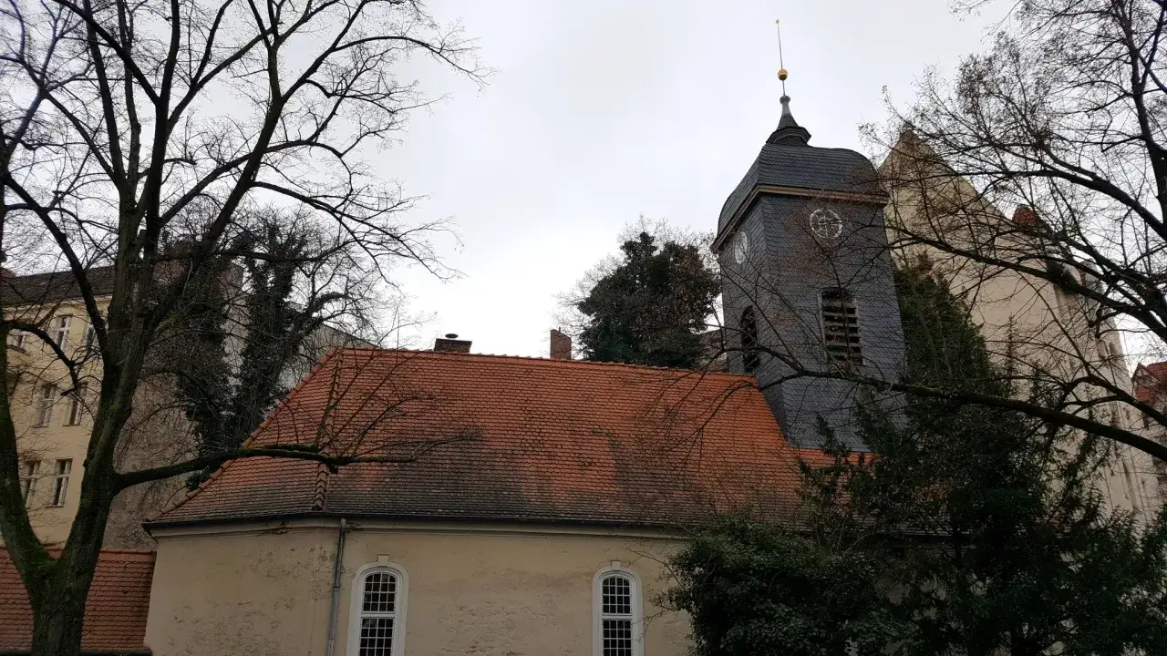 Kirchturm und rote Ziegeld&auml;cher in Rixdorf, Berlin, umrahmt von kahlen B&auml;umen.