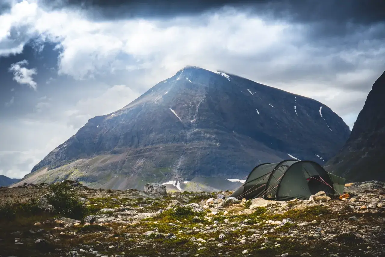 Tältläger vid foten av ett berg, perfekt för guidade turer i den svenska fjällvärlden. Dramatiska moln hänger över toppen.