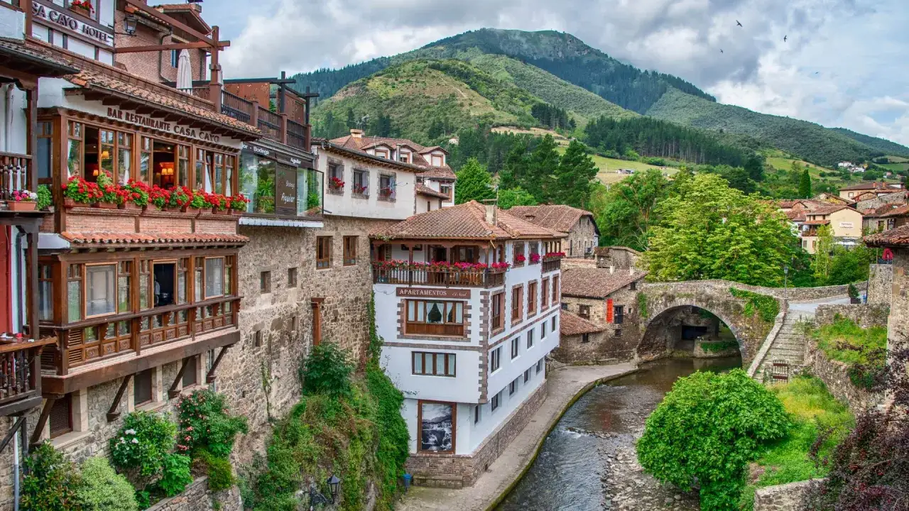 Pueblos costeros de Cantabria: casas de piedra y madera con balcones floridos junto a un río, bajo montañas verdes.