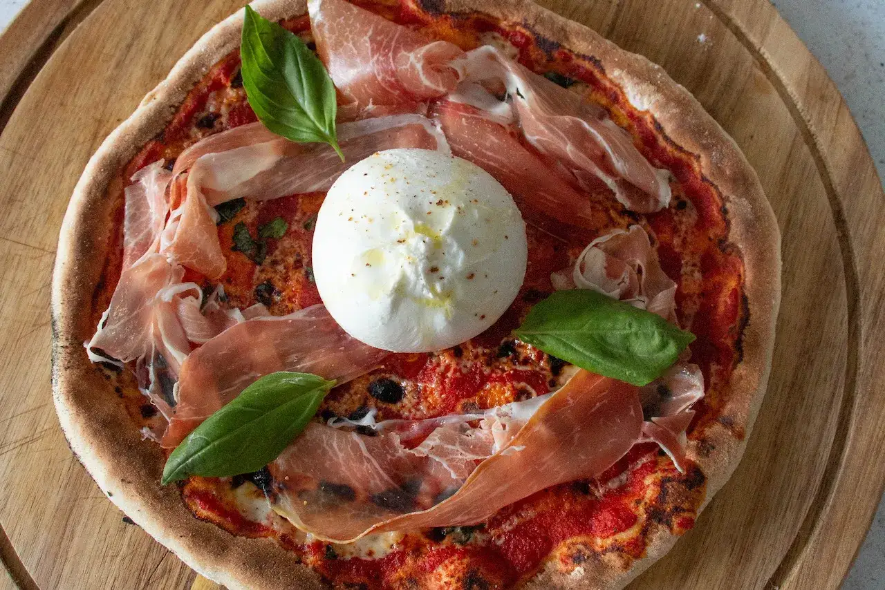 Pizza gourmande avec une burrata crémeuse au centre, garnie de fines tranches de prosciutto et de feuilles de basilic frais. Un délice !