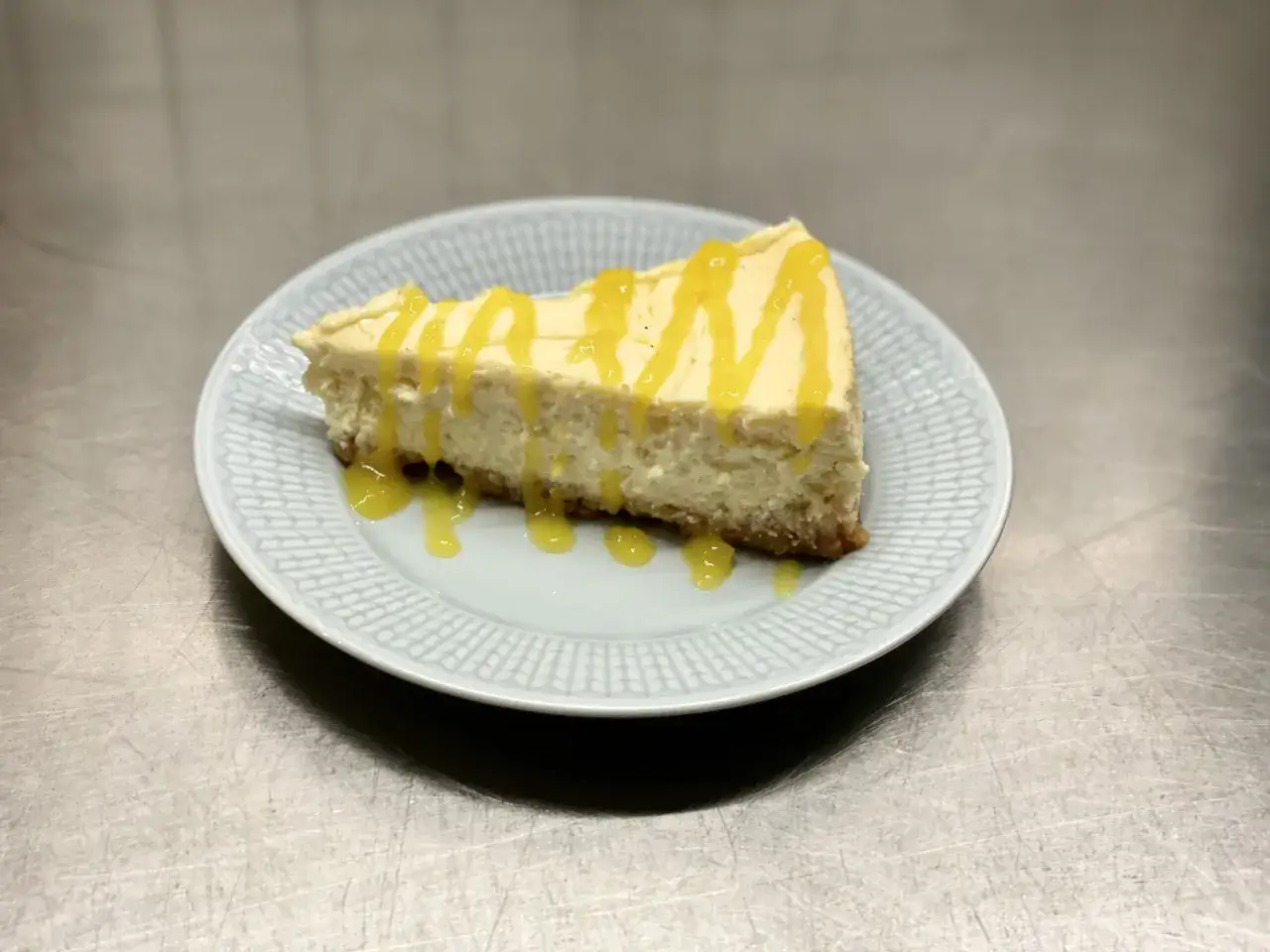 En bit kr&auml;mig mango cheesecake med gul glasyr p&aring; en ljusbl&aring; tallrik.