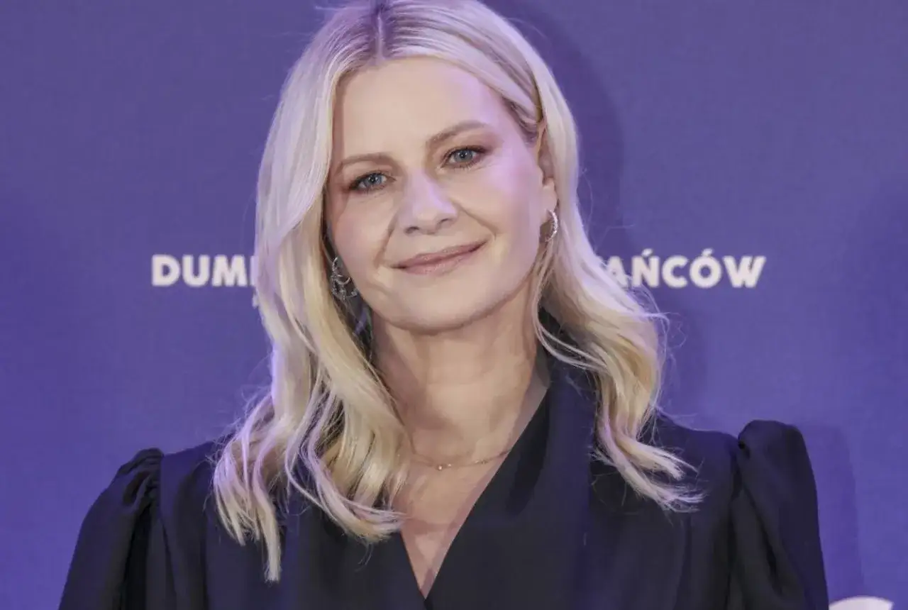 Uśmiechnięta blondynka z delikatnym makijażem i kolczykami, w czarnej bluzce z bufiastymi rękawami. Jej fryzura, clavi cut, dodaje jej elegancji.