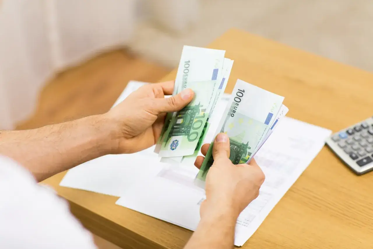 Dłonie liczą banknoty 100 euro. Obok leży kalkulator i dokumenty, sugerujące rozliczenie wynagrodzenia postojowego.