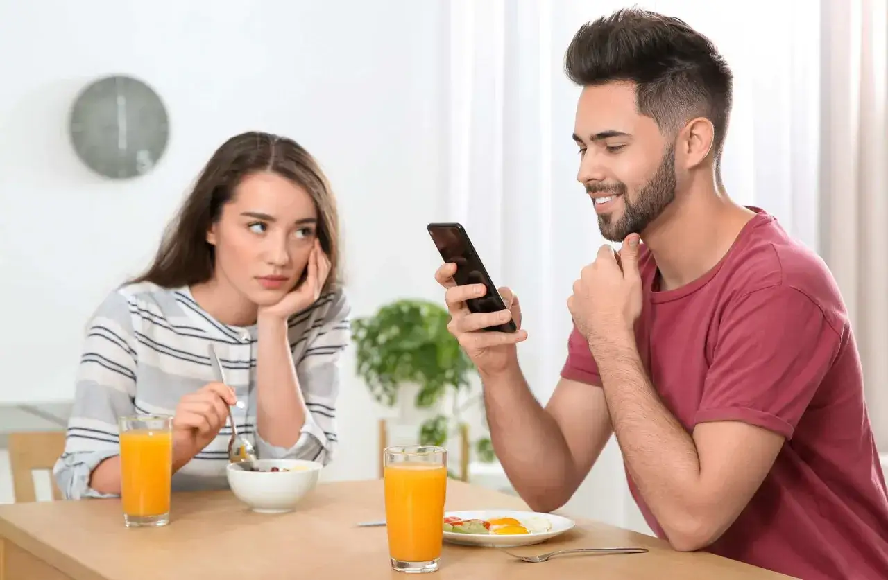 Smutna dziewczyna przy śniadaniu, podczas gdy chłopak ignoruje ją, przeglądając telefon. Czy to ghosting co to znaczy?