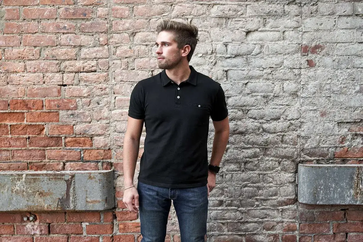 Uomo con un outfit con polo uomo nera e jeans, appoggiato a un muro di mattoni.