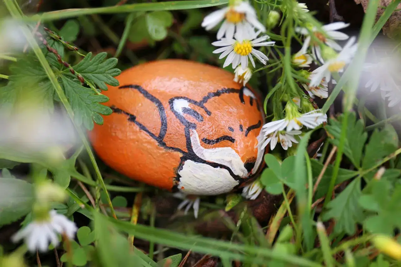 Bemalte Steine: Ein orangefarbener Stein mit einem Fuchsgesicht liegt im Gras, umgeben von kleinen wei&szlig;en Blumen und gr&uuml;nen Bl&auml;ttern.