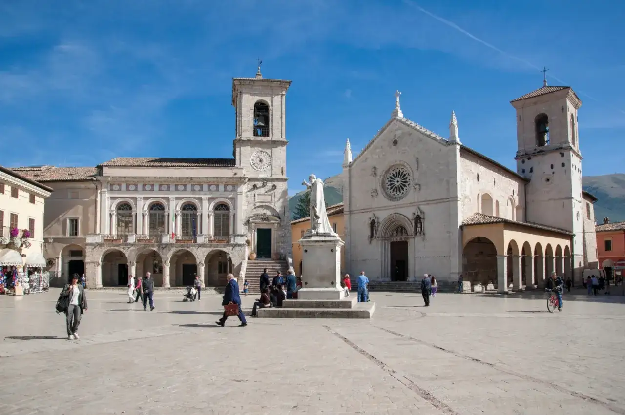 A sunny day in Nursia's main piazza, with a statue, a clock tower, and a church.