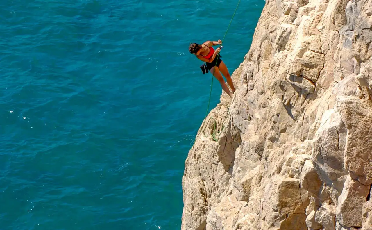 Frau seilt sich an einer Felswand ab, das Meer glitzert türkisblau. Ein Abenteuer beim Finale Ligure Klettern.