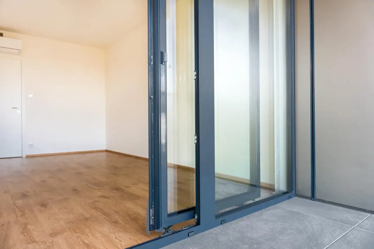 Detalle ventana corredera gris con marcos finos y cristalera, que da a una habitación vacía con suelo de madera.