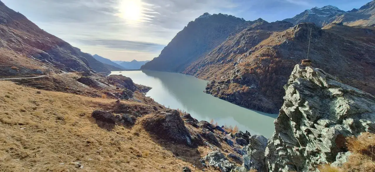 Paesaggio alpino con lago artificiale e montagne scoscese. Il ghiacciaio Fellaria si riflette nelle acque calme, circondato da erba secca e rocce.