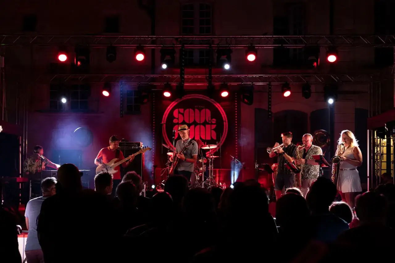 Un groupe de musique joue sur sc&egrave;ne, &eacute;clair&eacute; par des projecteurs rouges. Un chanteur soul fran&ccedil;ais, casquette sur la t&ecirc;te, joue de la guitare.
