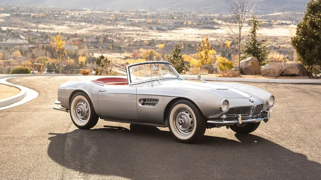 Srebrny kabriolet BMW 507 z czerwonym wnętrzem, zaparkowany na podjeździe z widokiem na jesienny krajobraz.