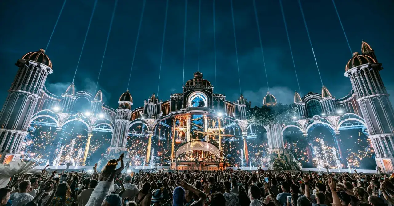 La folla esulta sotto un'imponente scenografia architettonica illuminata da fuochi d'artificio e fasci di luce, un'emozionante anteprima del line up Tomorrowland.