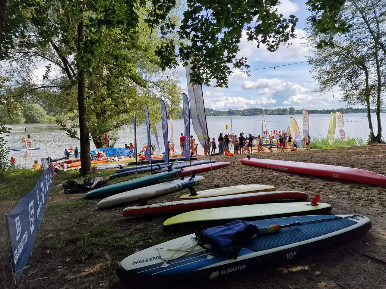 Deska SUP na piasku nad jeziorem Przeczyckim, w tle ludzie i sprzęt wodny.