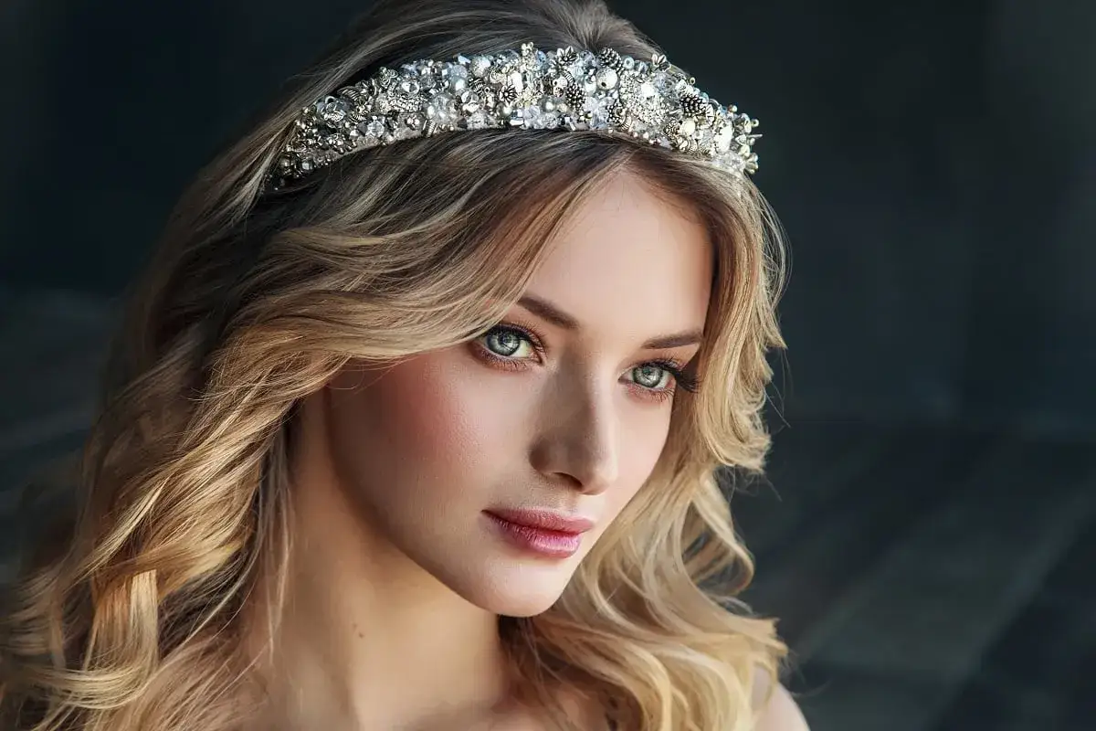 Piękna kobieta z falowanymi blond włosami nosi bogato zdobioną tiarę srebrną, która dodaje jej królewskiego blasku.