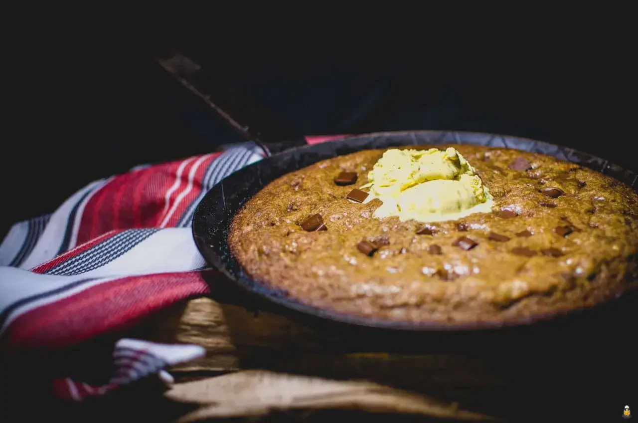 Ein warmer, schokoladiger Skillet Cookie mit einer Kugel Vanilleeis, serviert in einer Gusseisenpfanne, daneben ein rot-weiß gestreiftes Tuch.